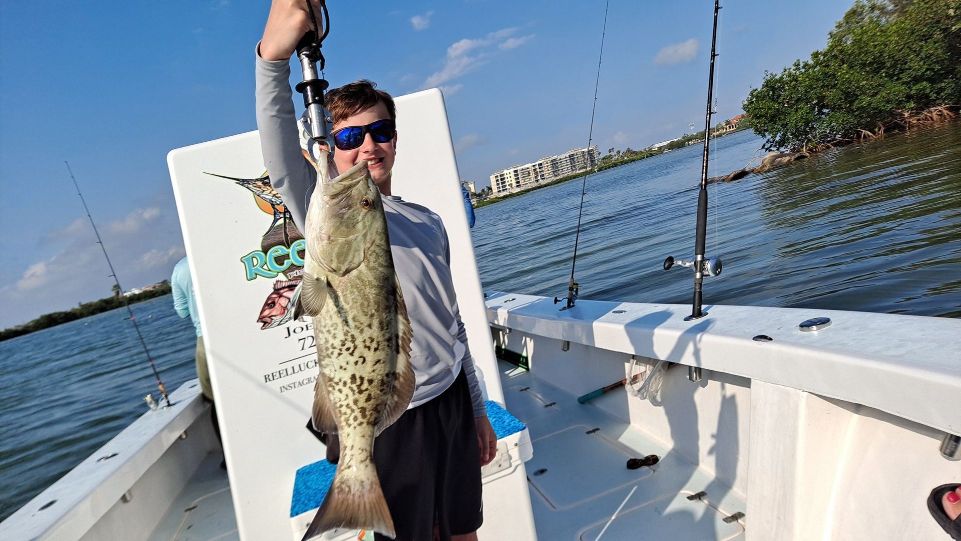 Young man on a boat holds up a speckled fish, blue sky, and water.