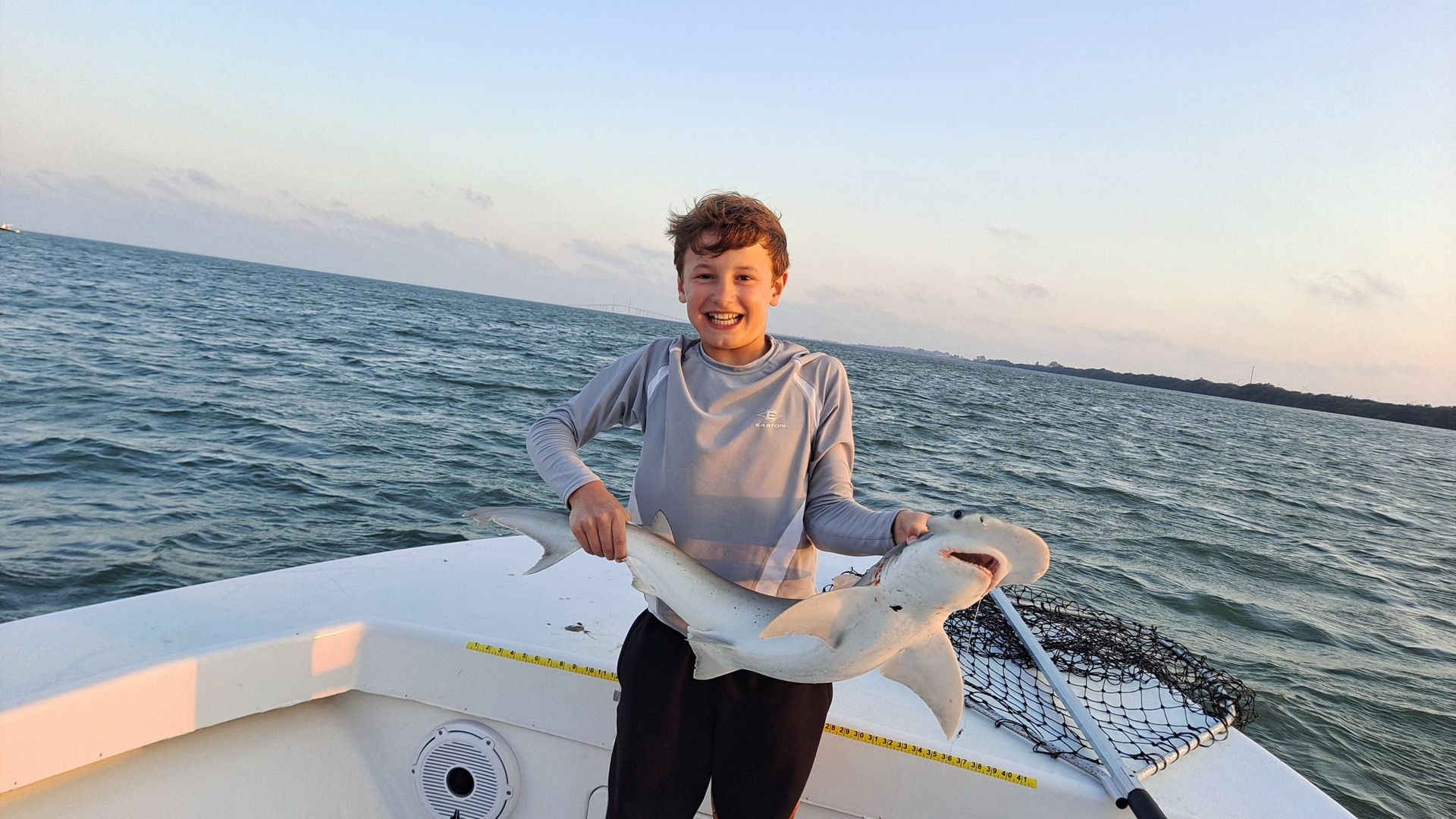 Boy on boat holds up small shark, smiling, with ocean background.