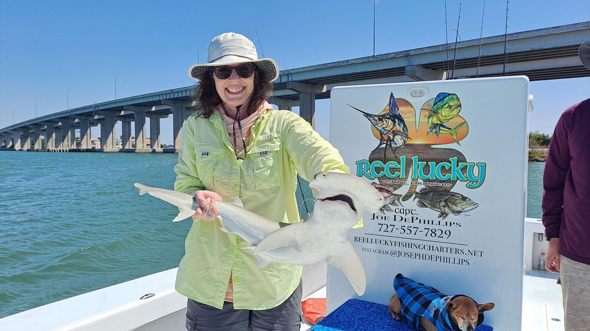 Woman smiling, holding a small hammerhead shark on a boat, bridge in background.