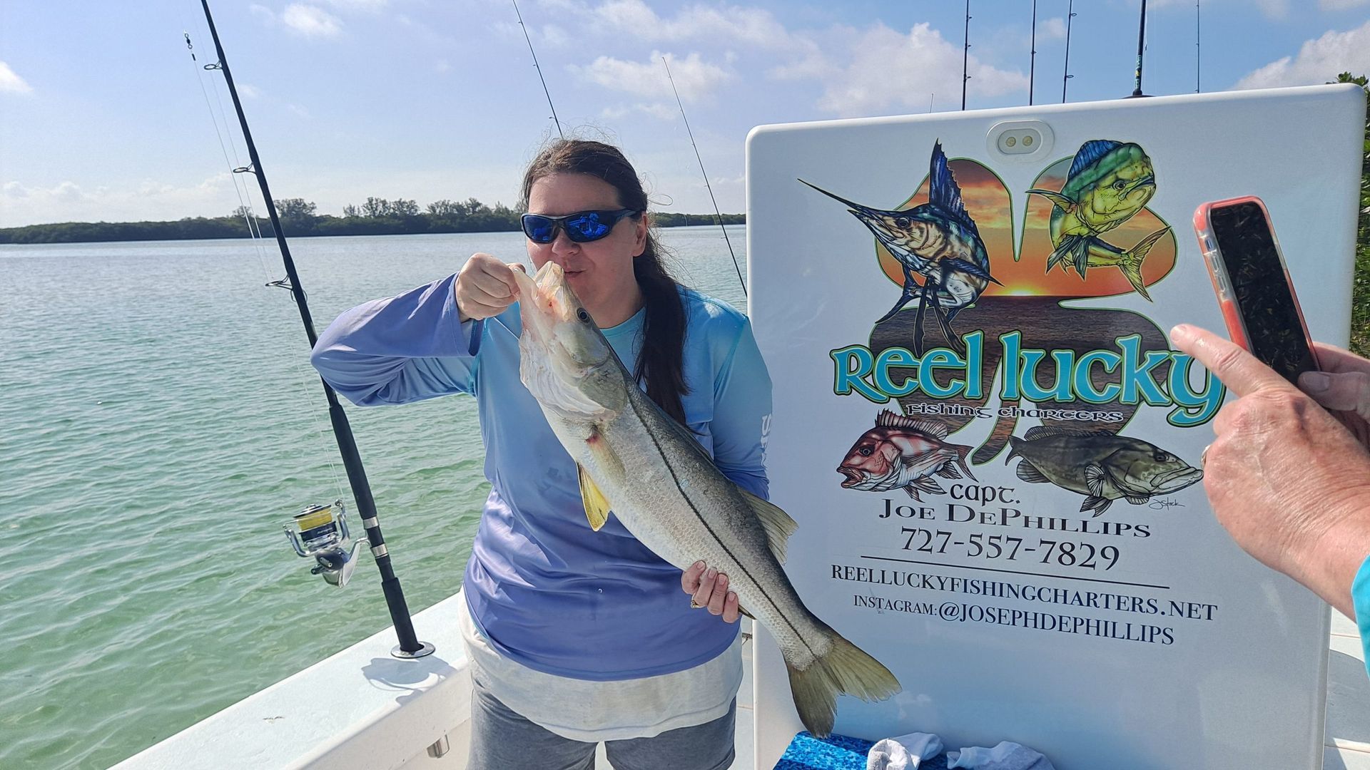 Woman on a boat holds a caught fish, Reel Lucky sign behind her. Blue skies and water.