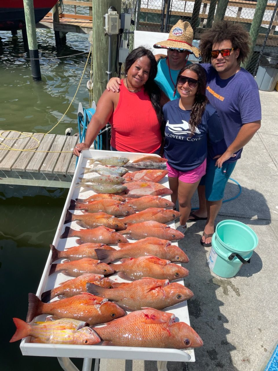 Four people pose with a table of caught red fish at a dock. Sunny day.