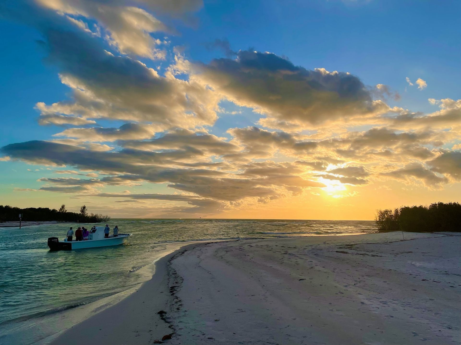 Boat on a white sand beach at sunset with people on board. Orange sun glows through clouds.