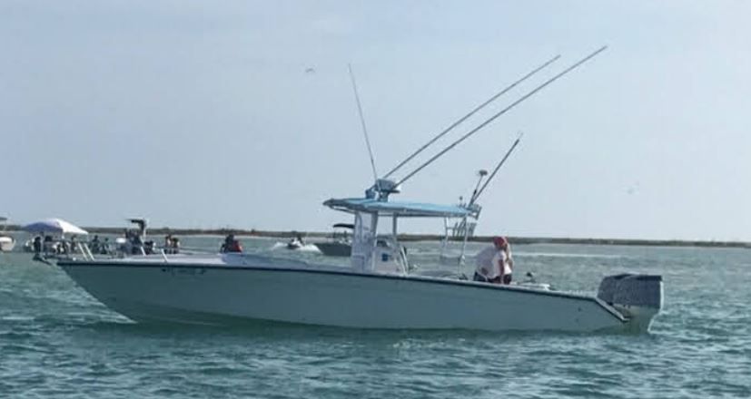 A light blue fishing boat on water with fishing rods, a man at the helm, and the ocean with a hazy sky.