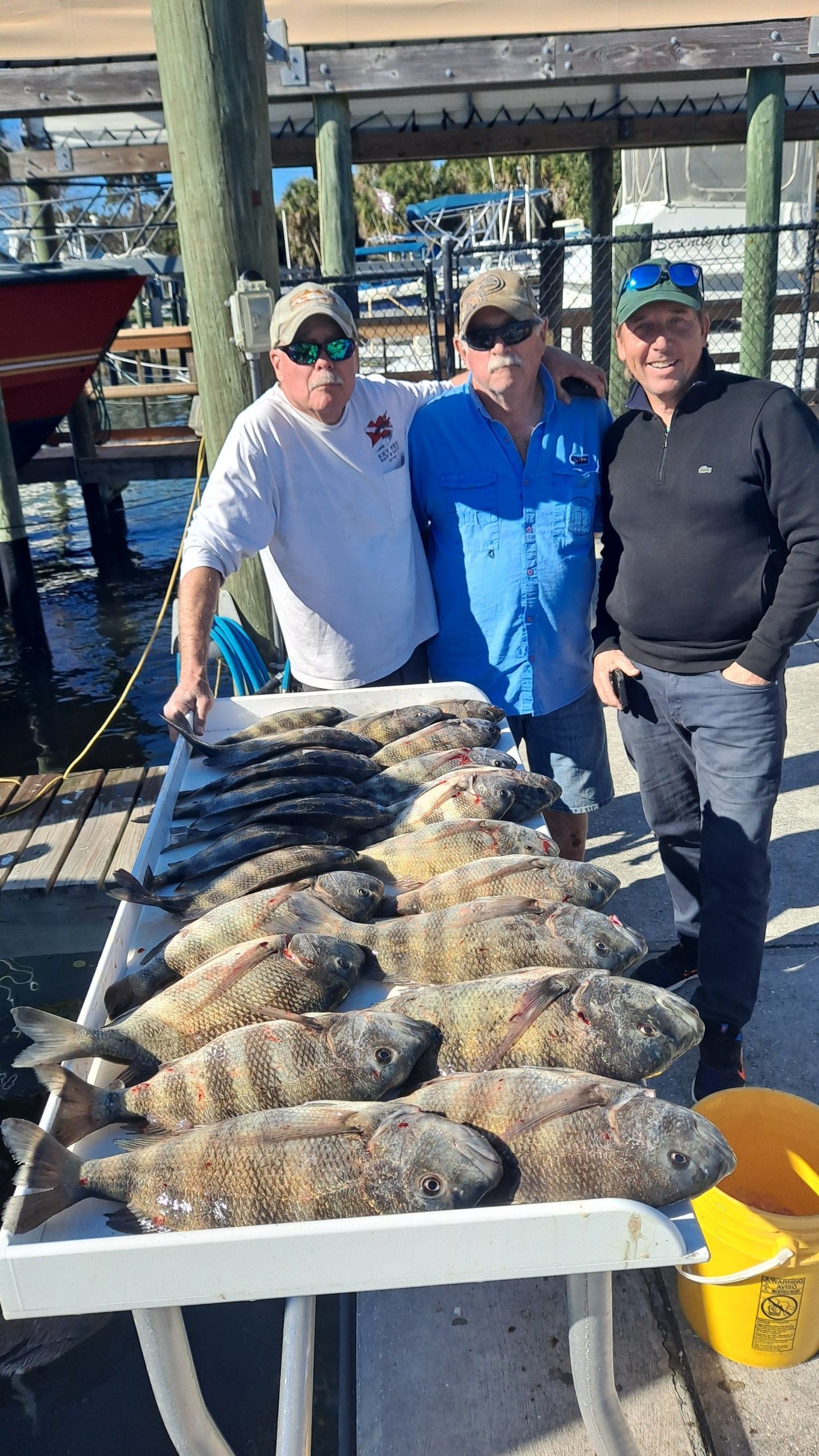 Three men pose with a table of caught fish at a dock. Cloudy sky.