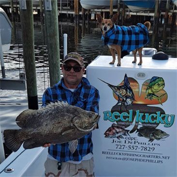 Man holds a large fish on a boat, a dog in a coat stands nearby. The logo says 