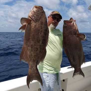 Man on a boat holds two large, dark fish, blue sea in background.