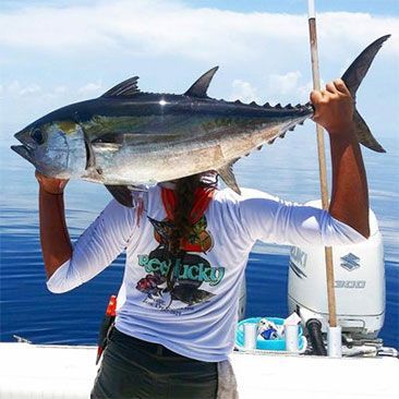 Person holding a large tuna fish over their head on a boat. Blue water and sky in background.