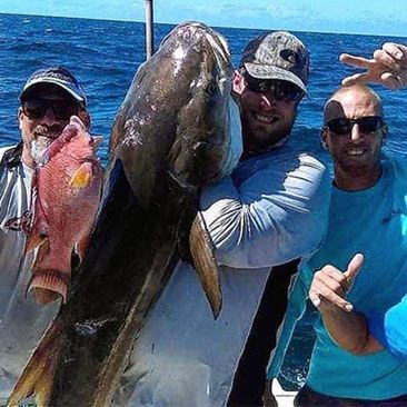 Three men on a boat hold large fish in the ocean, one large brown fish and one red fish; all smiling.