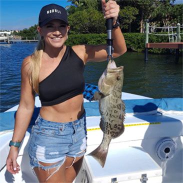 Woman on boat, holding a fish she caught. Wearing shorts and a black top, smiling.