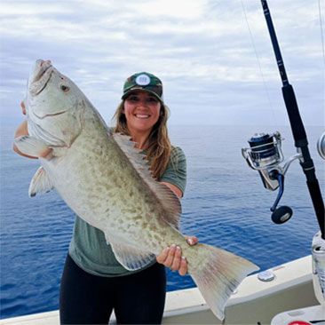 Woman on boat smiles holding a large fish, blue water background.