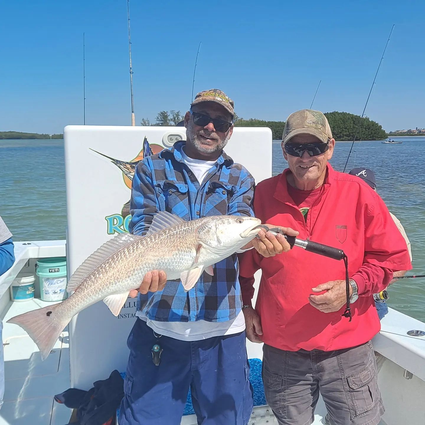 Two men on a boat display a large redfish. Clear sky, sunny day. One man holds fish, other smiles.