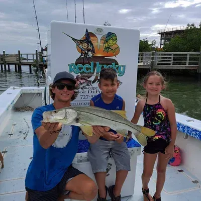 Three people on a boat holding a snook. Man kneeling, two children standing, all smiling. Fishing boat in the background.