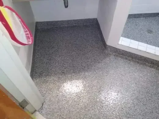 Bathroom corner with speckled gray floor, white walls, and shower entrance. A bag hangs on the wall.