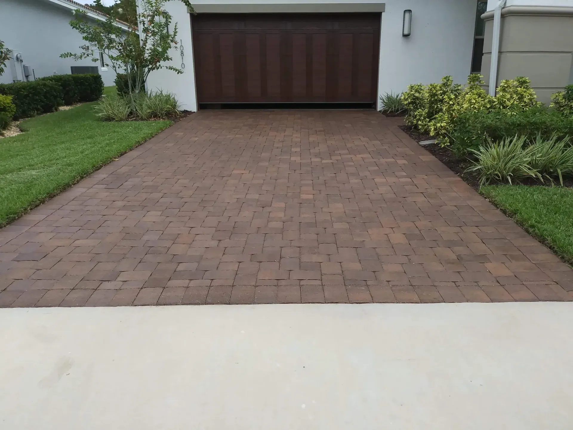 Brick driveway leading to a brown garage door, flanked by green grass and shrubbery.