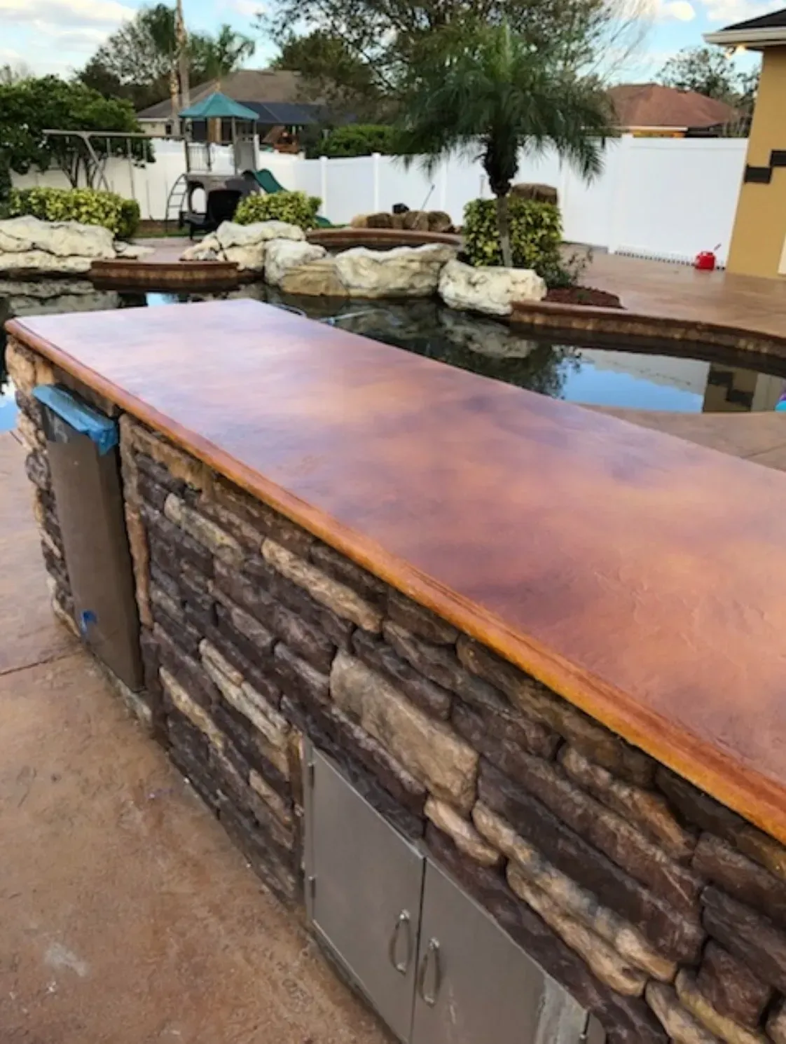 Outdoor kitchen with brown stone, a countertop, and a pool in the background.