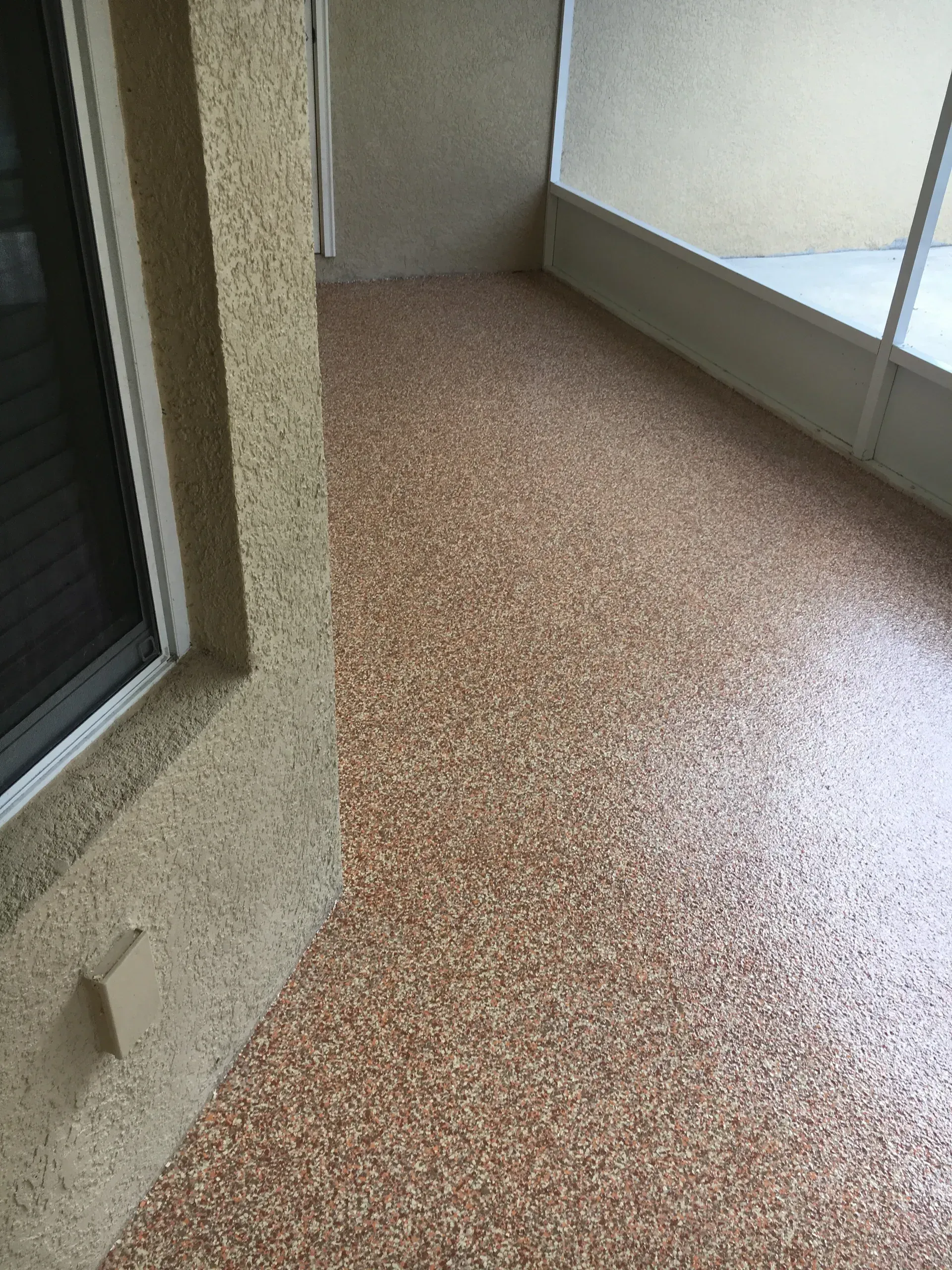 A porch with a speckled brown floor, beige walls, and a window on the left side.