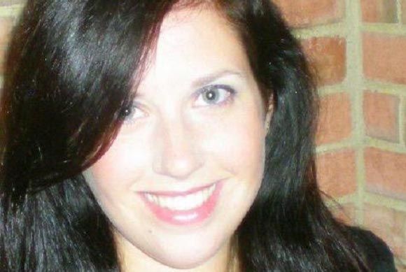 A woman with long dark hair is smiling in front of a brick wall.