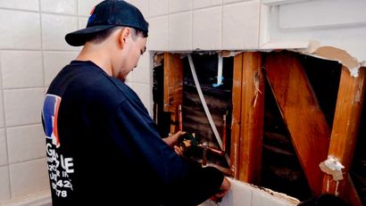 Person in a black cap and shirt works on exposed wall framing inside a damaged room.