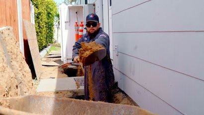 Worker in a narrow trench pours dirt beside a white building wall.