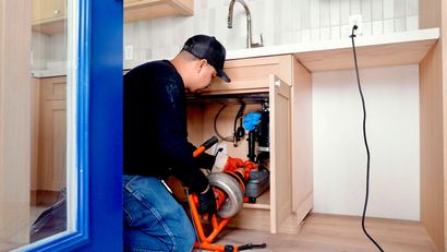 Plumber working under a kitchen sink with tools and exposed pipes, beside a light wood cabinet.