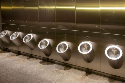 Row of stainless steel urinals on a tiled restroom wall, each lit by bright circular lights.
