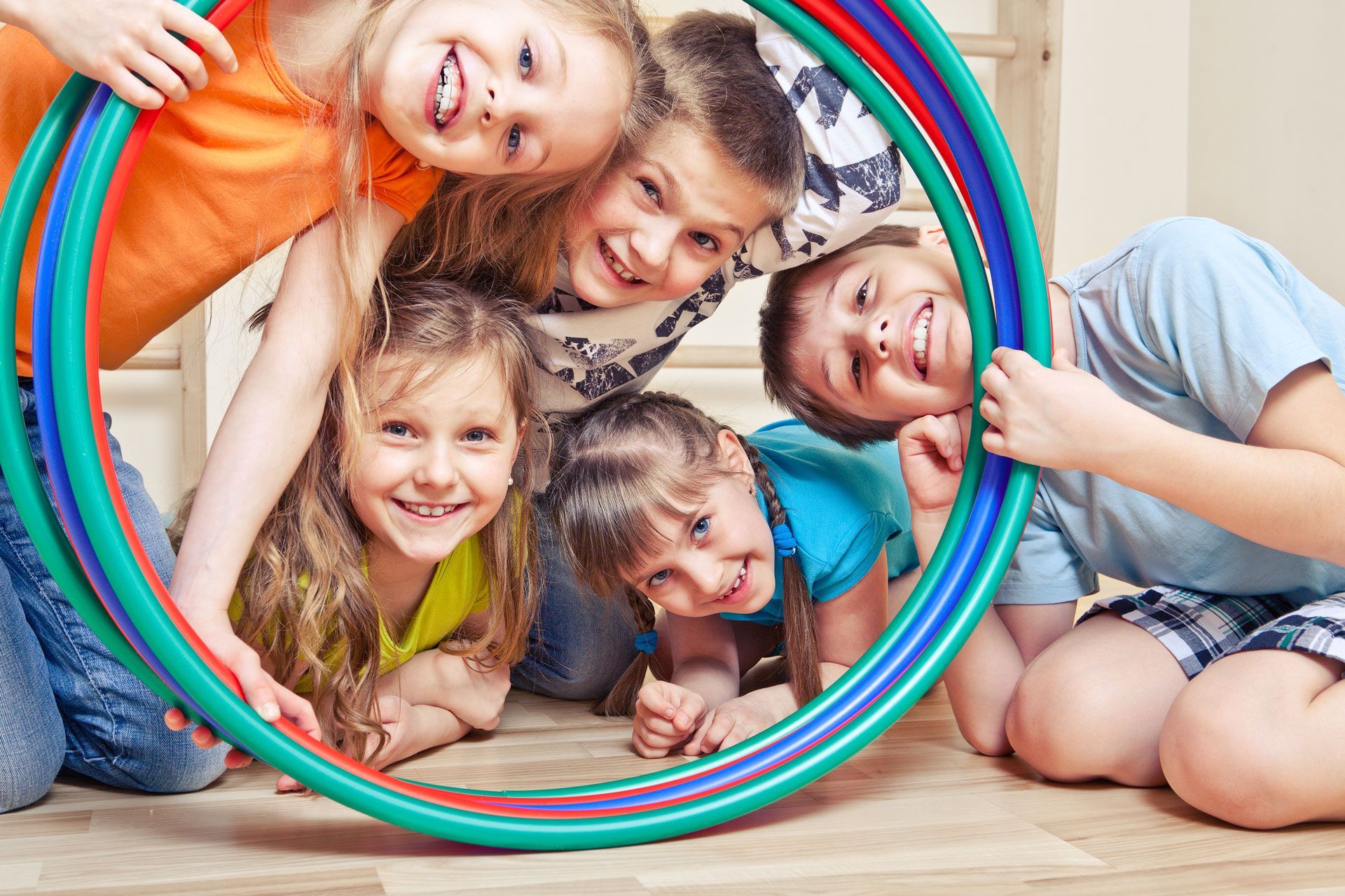 Children smiling, looking through colorful hoops on a wooden floor.