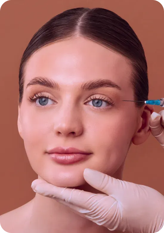 Woman receiving an injection near her eye; a gloved hand holds her face, brown background.