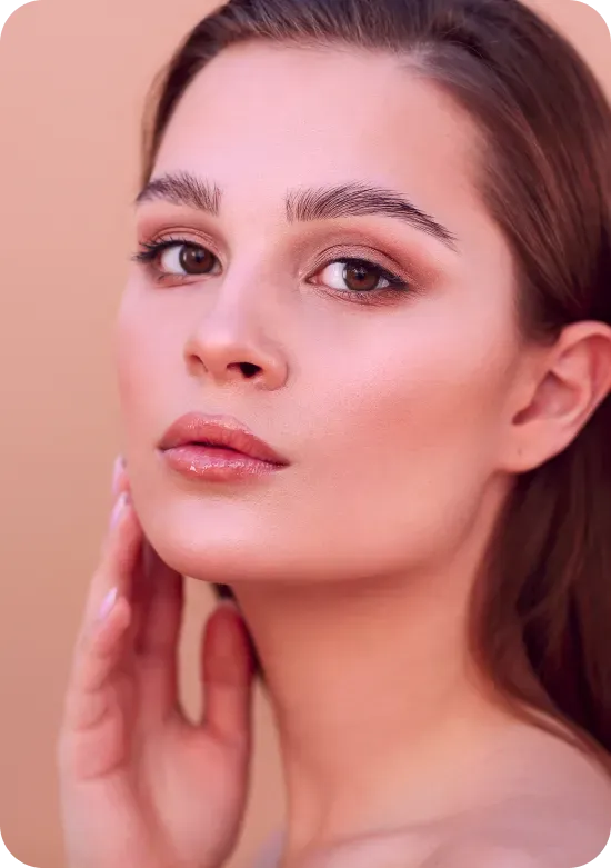 Woman with brown hair and light makeup poses, hand touching face, against a peach background.