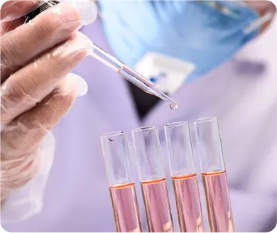 Scientist's gloved hand with dropper adding liquid to test tubes containing pink liquid in a lab setting.