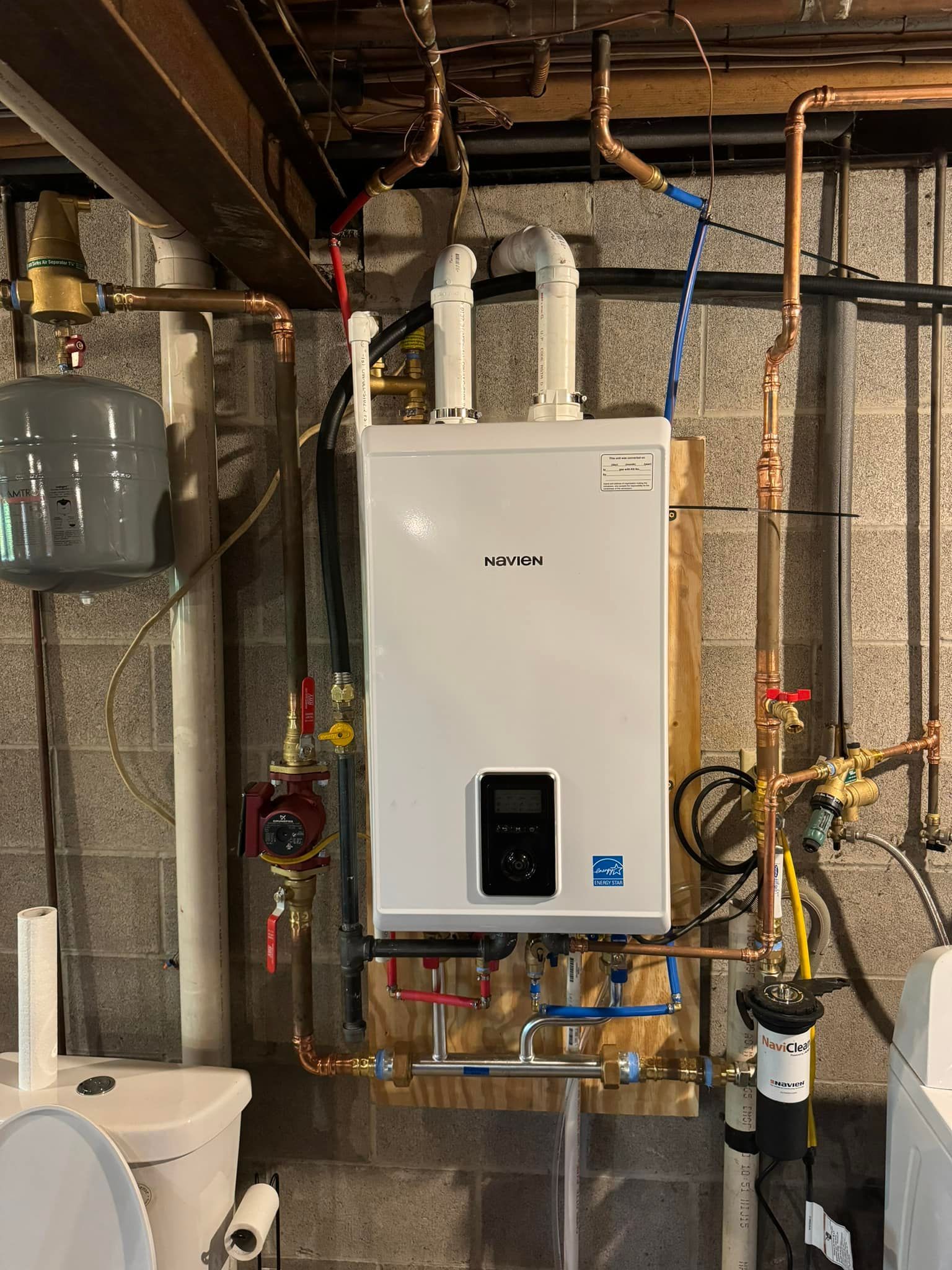 A white wall-mounted boiler with copper pipes against a concrete block wall in a basement setting.