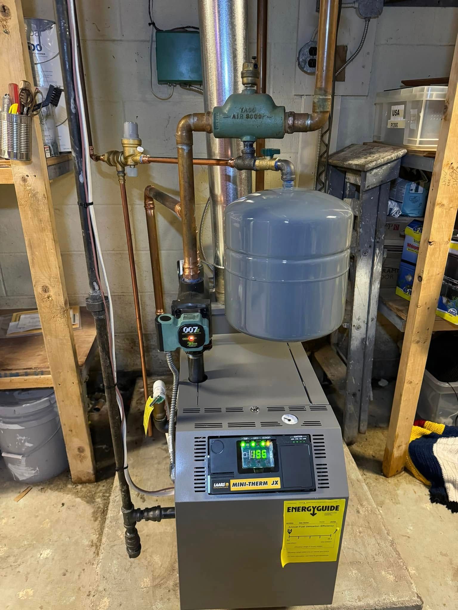 A gray boiler with copper pipes, a pressure tank, and digital display.  Located in a basement with wood shelving.