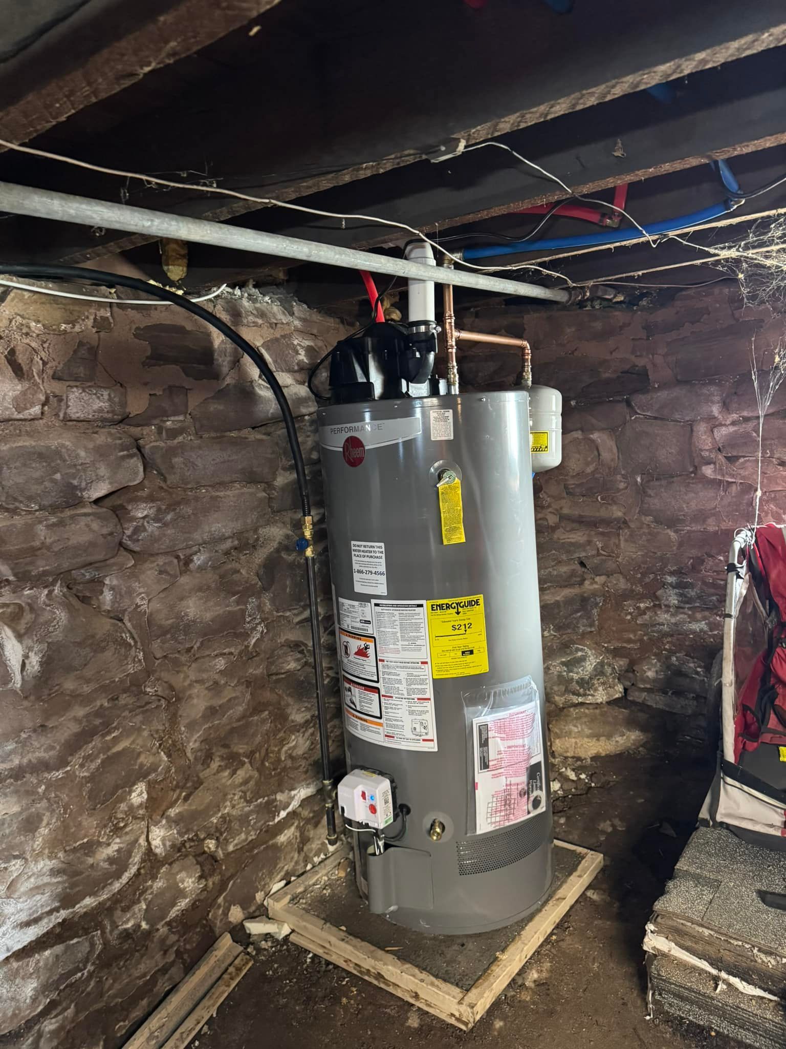 A vertical water heater in a basement. Grey tank, black top, on a wooden frame, against a stone wall.