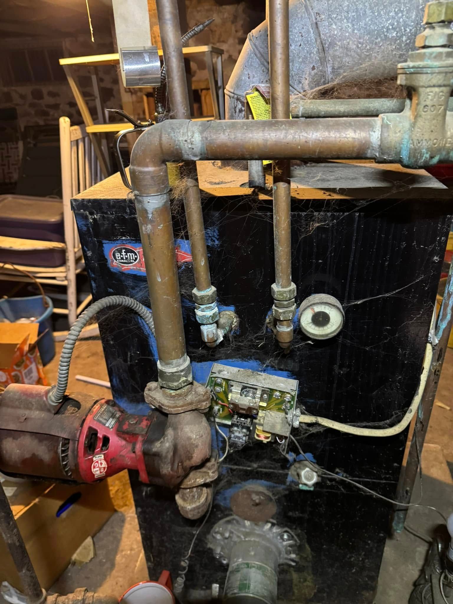 Black boiler with copper pipes and red pump. Indoors, with wood and other equipment visible.