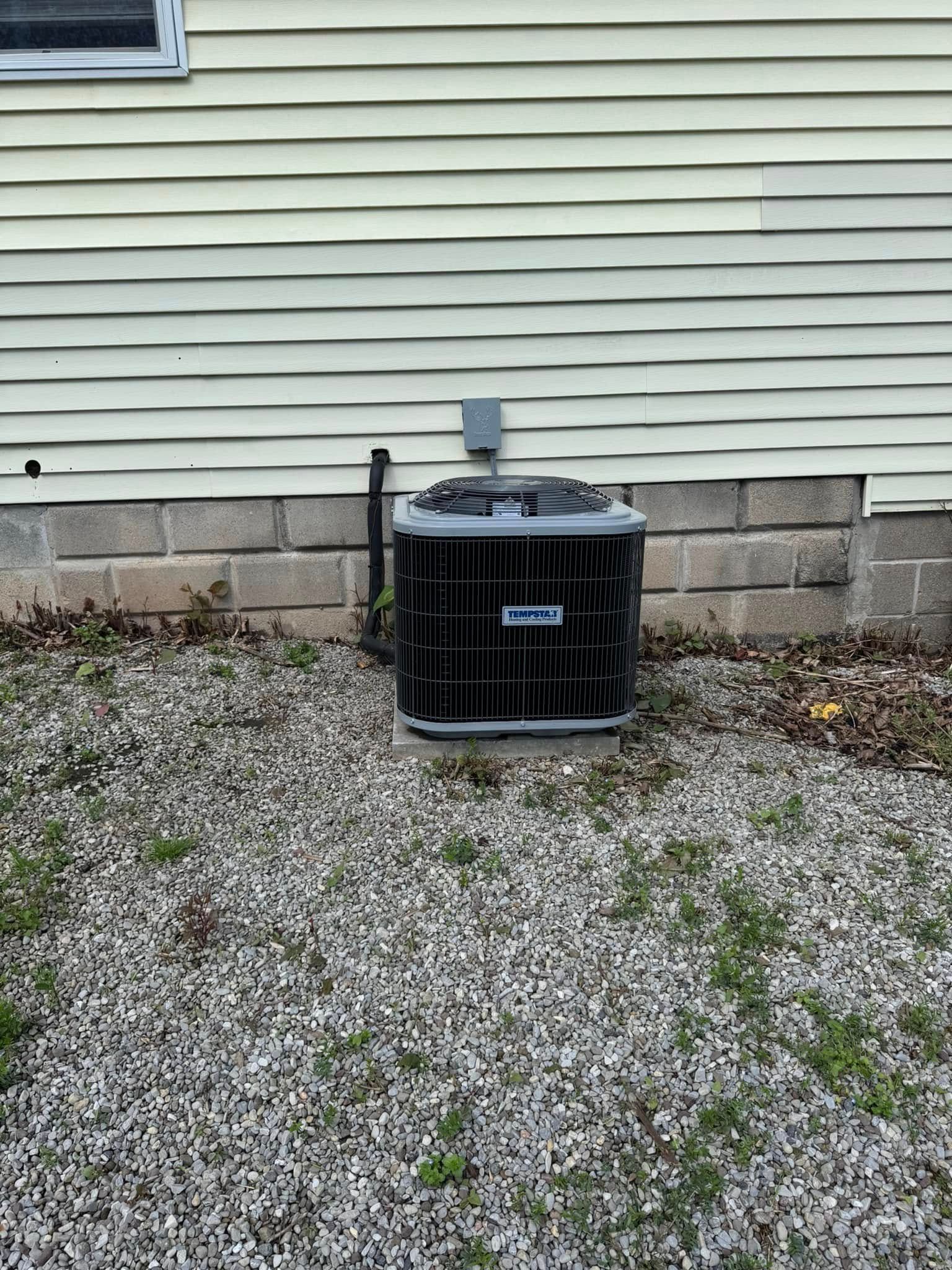 Black HVAC unit on gravel near a light yellow house with a concrete block foundation.