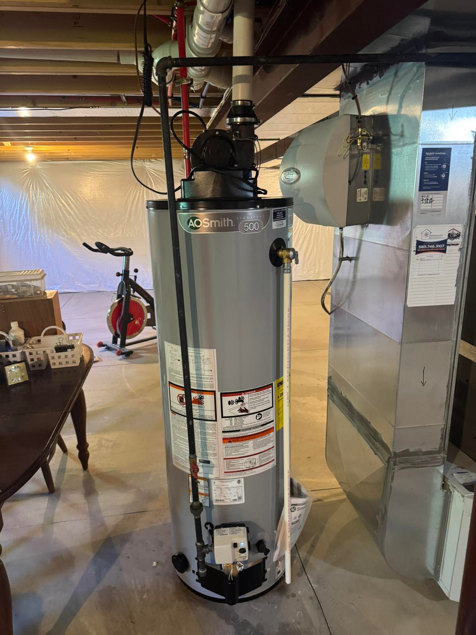 A basement with a tall water heater and HVAC unit, near a table, exercise bike, and plastic sheeting.