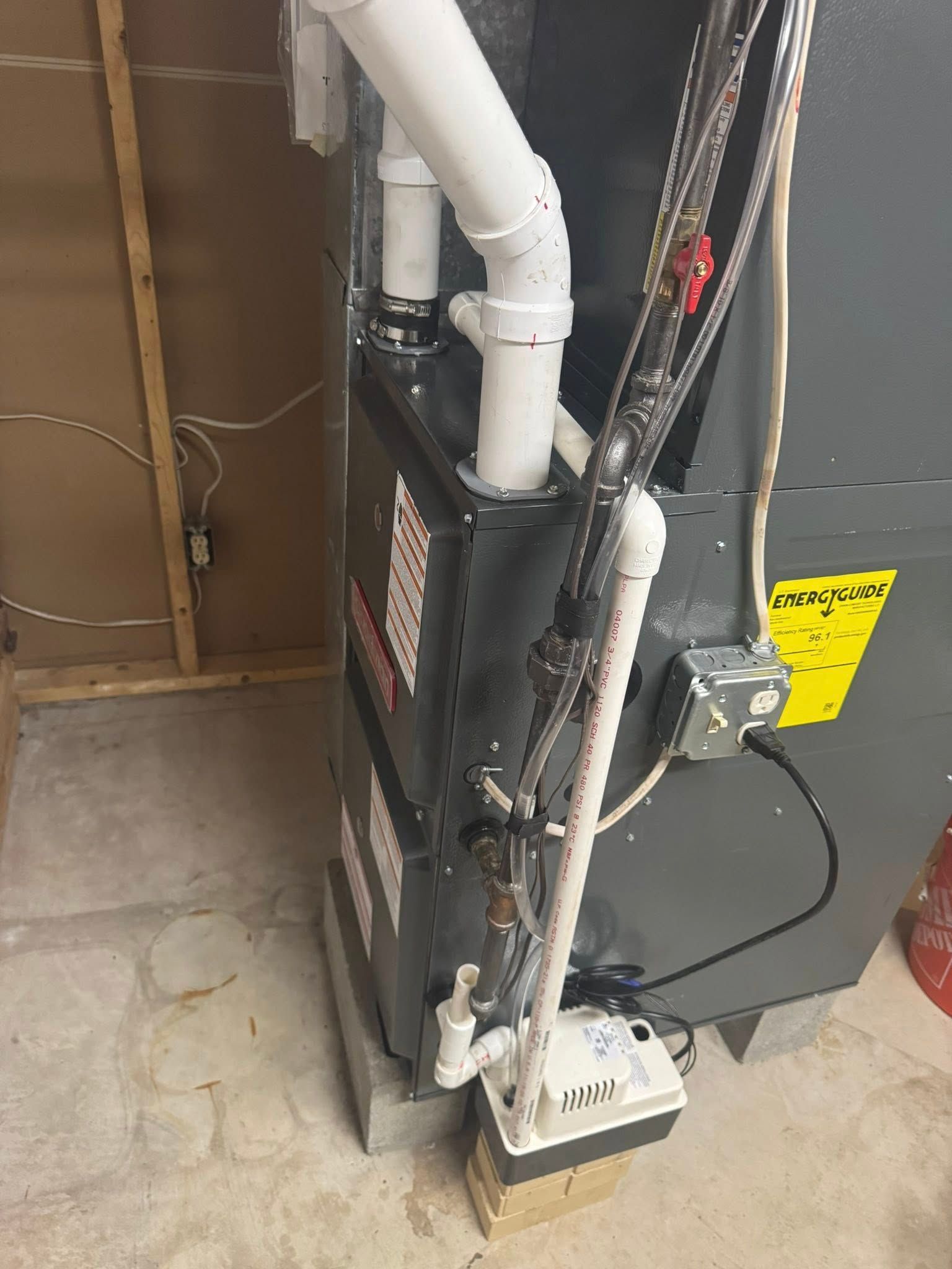 Furnace with white PVC pipes, electrical wiring, and a condensate pump, in a utility room.