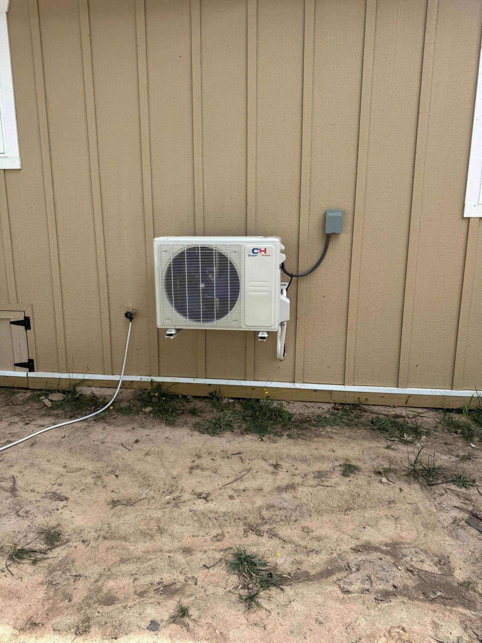 Beige outdoor AC unit mounted on a light brown wall. Electrical box and drainage pipe visible.