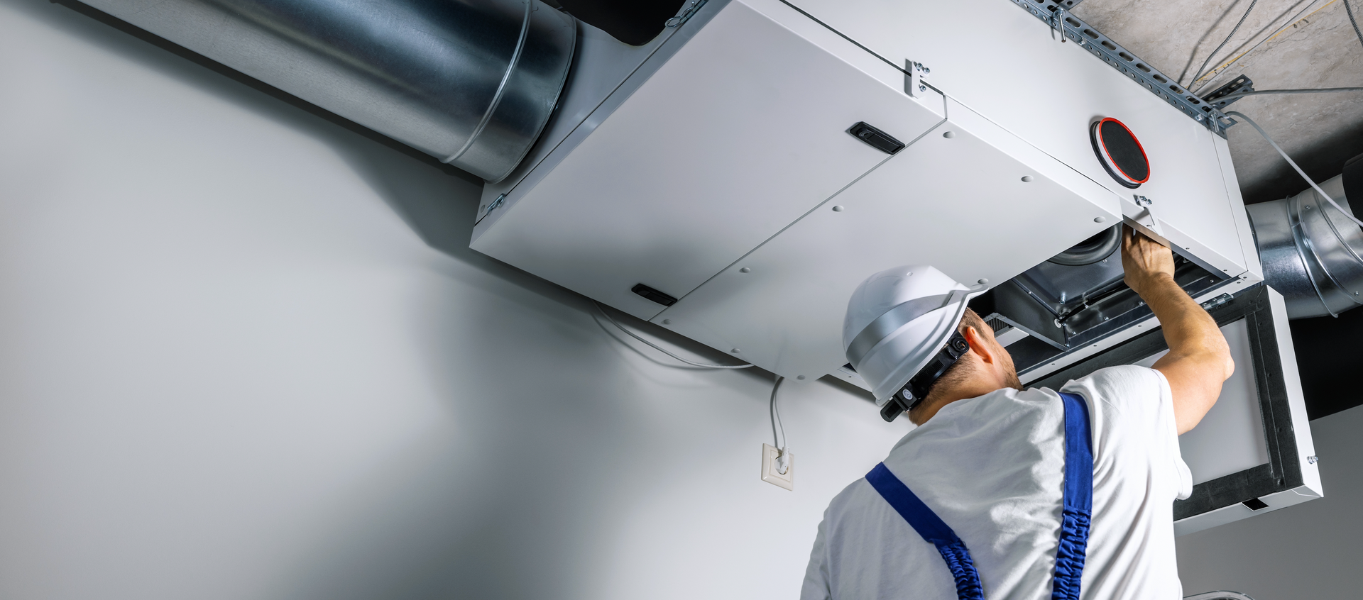 A technician in a white hard hat is installing a ventilation system in a white room.