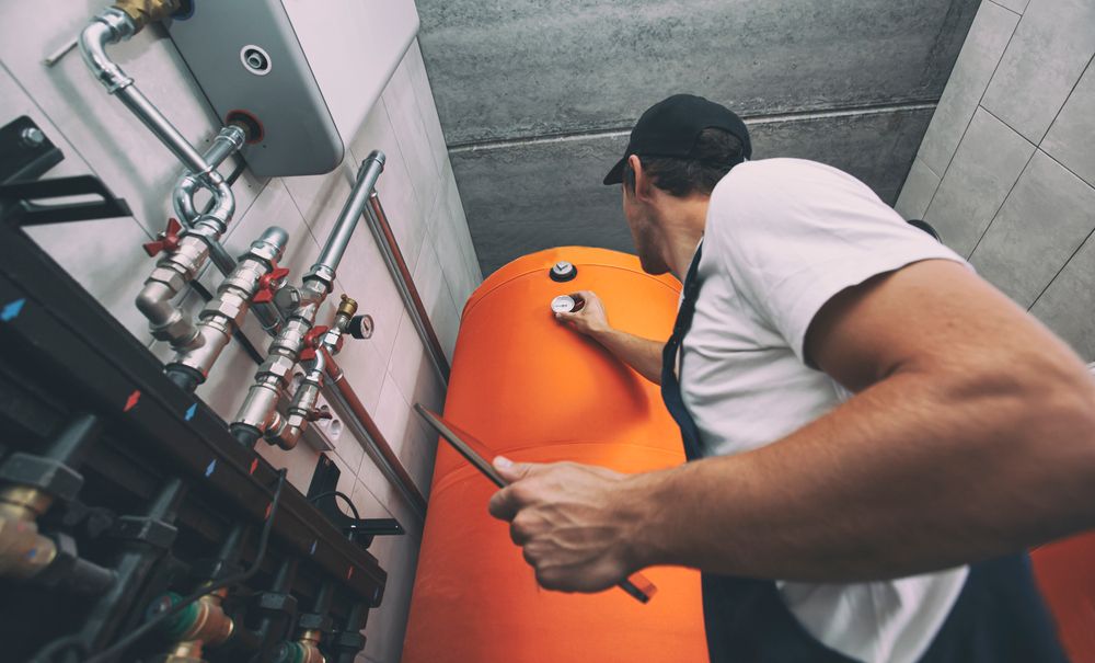 Plumber in a mechanical room inspecting a large orange water tank, checking gauges, holding a tool.