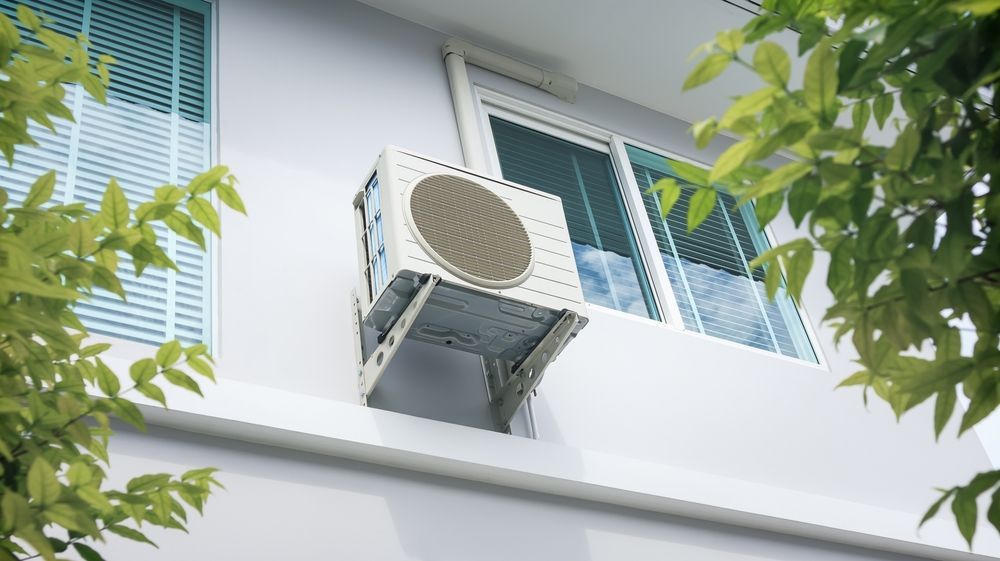 Air conditioner unit mounted on a white building exterior near a window, partly obscured by green leaves.