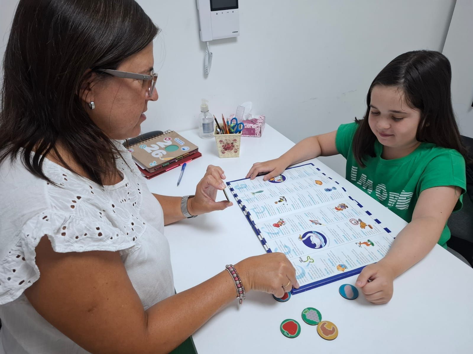 Una mujer y una niña juegan a un juego de mesa. La mujer señala el juego. La niña sonríe.