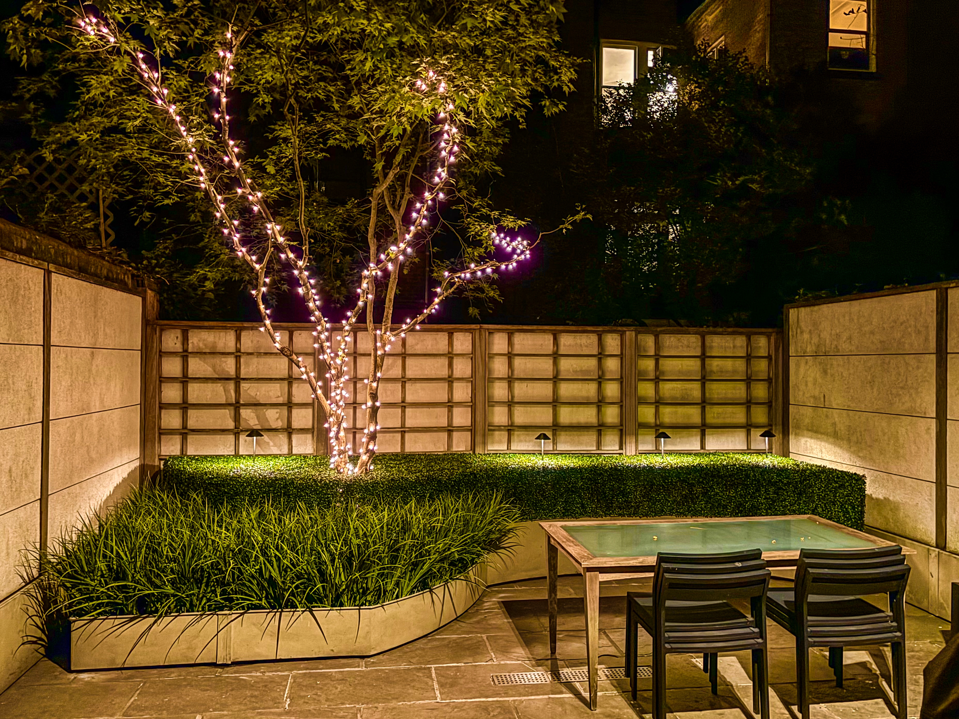 A patio area at night, featuring a small tree wrapped in pink fairy lights, a garden bench, and two outdoor chairs.