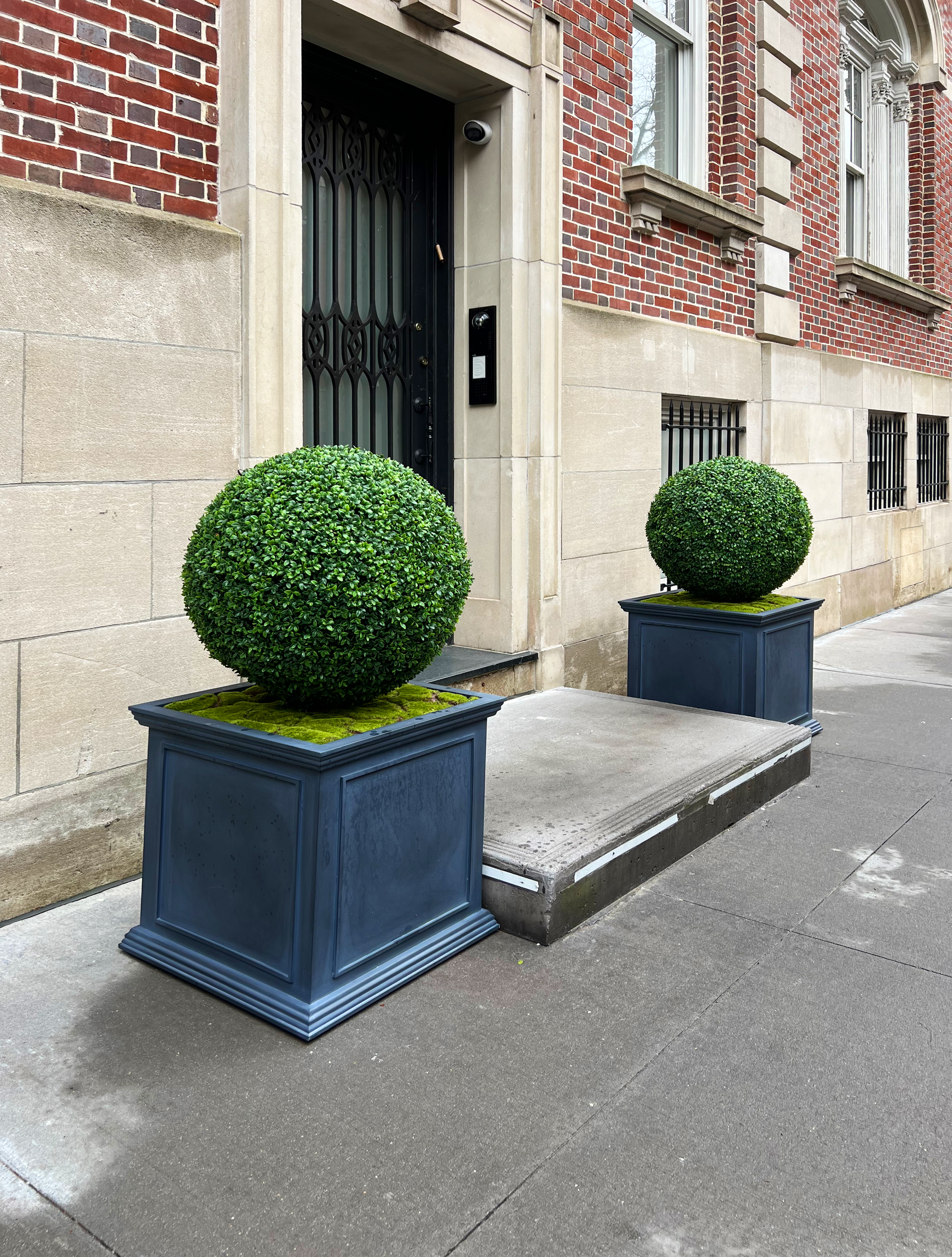 Two decorative spherical boxwood plants in dark blue square planters flank the entrance of a brick building.