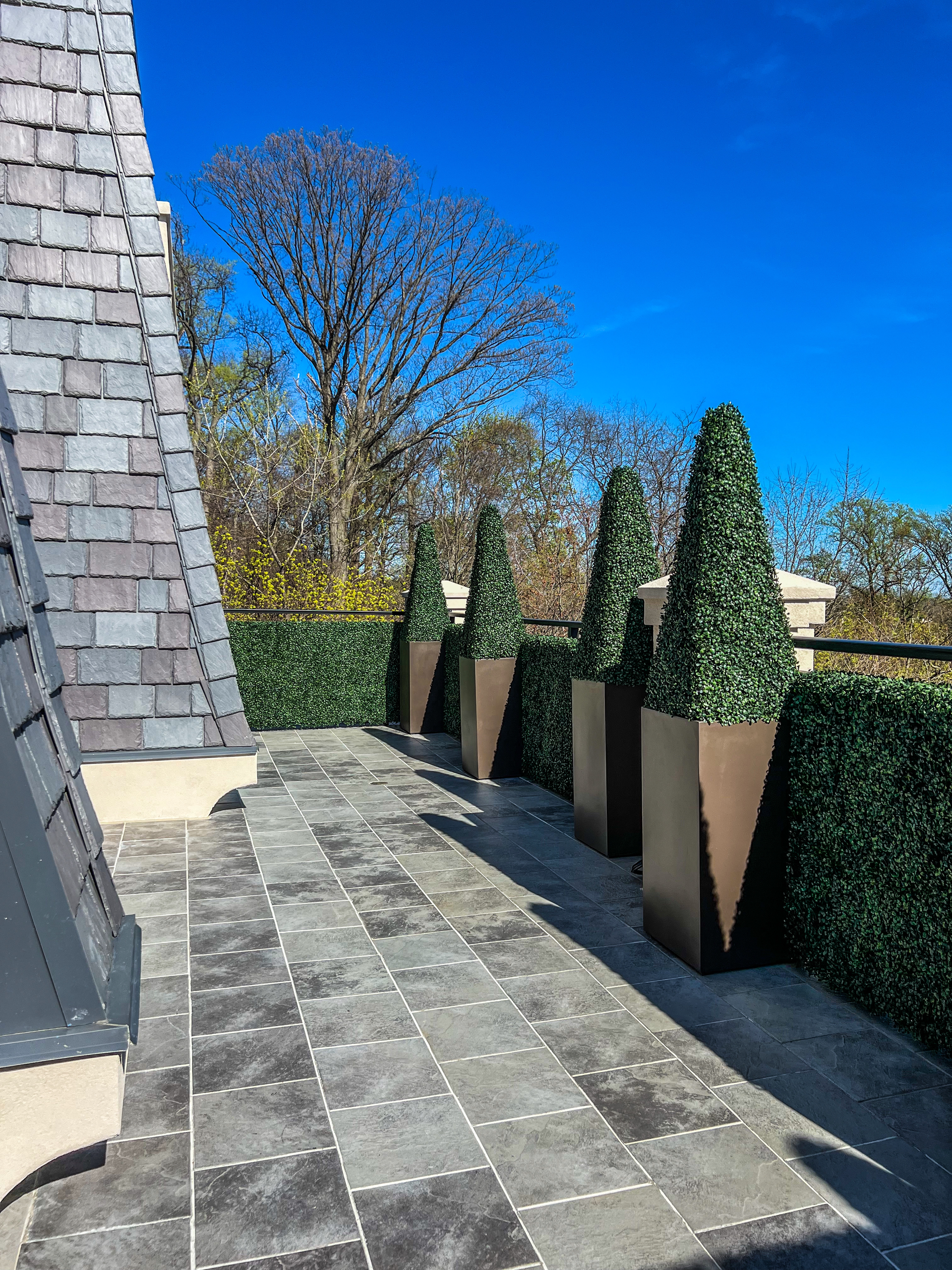 A stone-tiled terrace featuring four conical topiary trees in tall, brown planters against a green privacy hedge and sky.