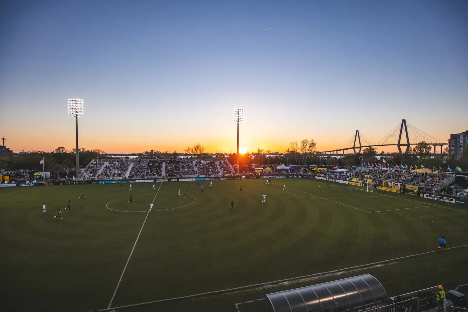 A soccer field with a crowd of people watching a game at sunset.