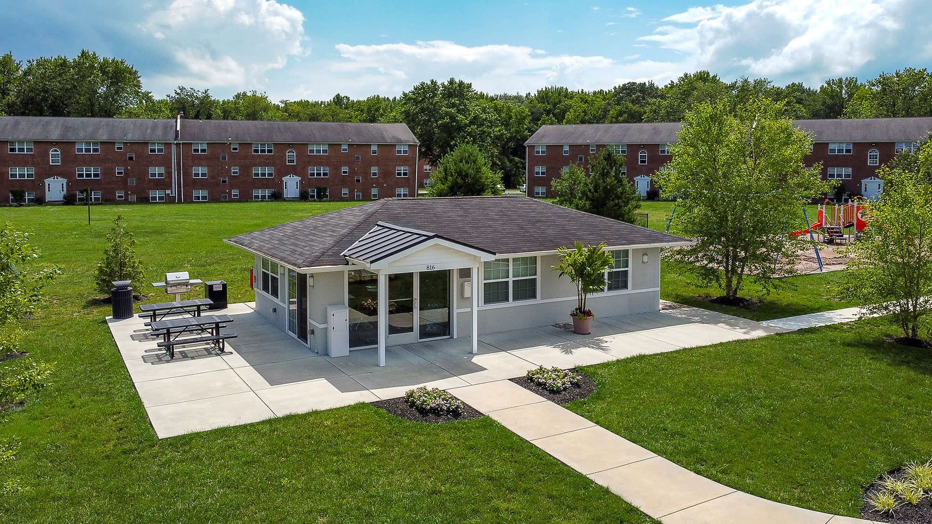 Single-story clubhouse with glass entry doors, concrete patio, and a nearby playground in a green apartment complex at Liberty Pointe in Newark, DE.