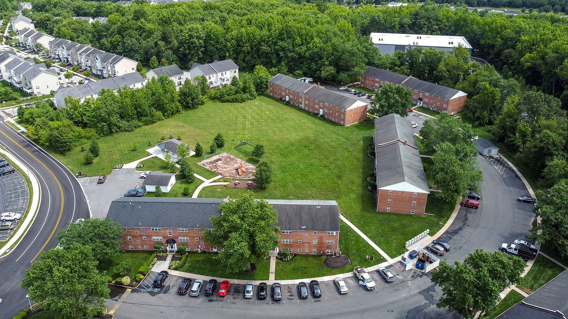Aerial view of a multifamily community with brick buildings, a large green lawn, playground, trees, and parking at Liberty Pointe in Newark, DE.