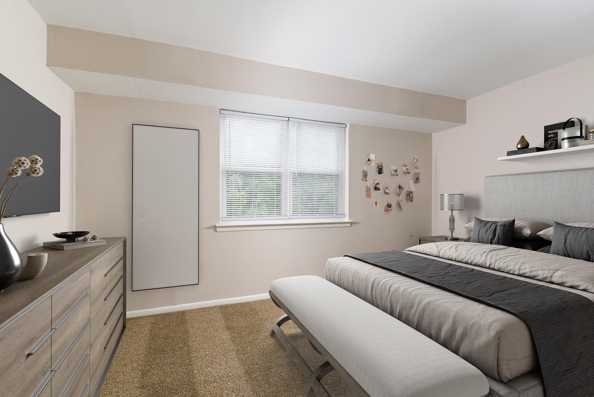 Bedroom in a modern apartment featuring a large bed, dresser, bench, and window with blinds at Liberty Pointe in Newark, DE.