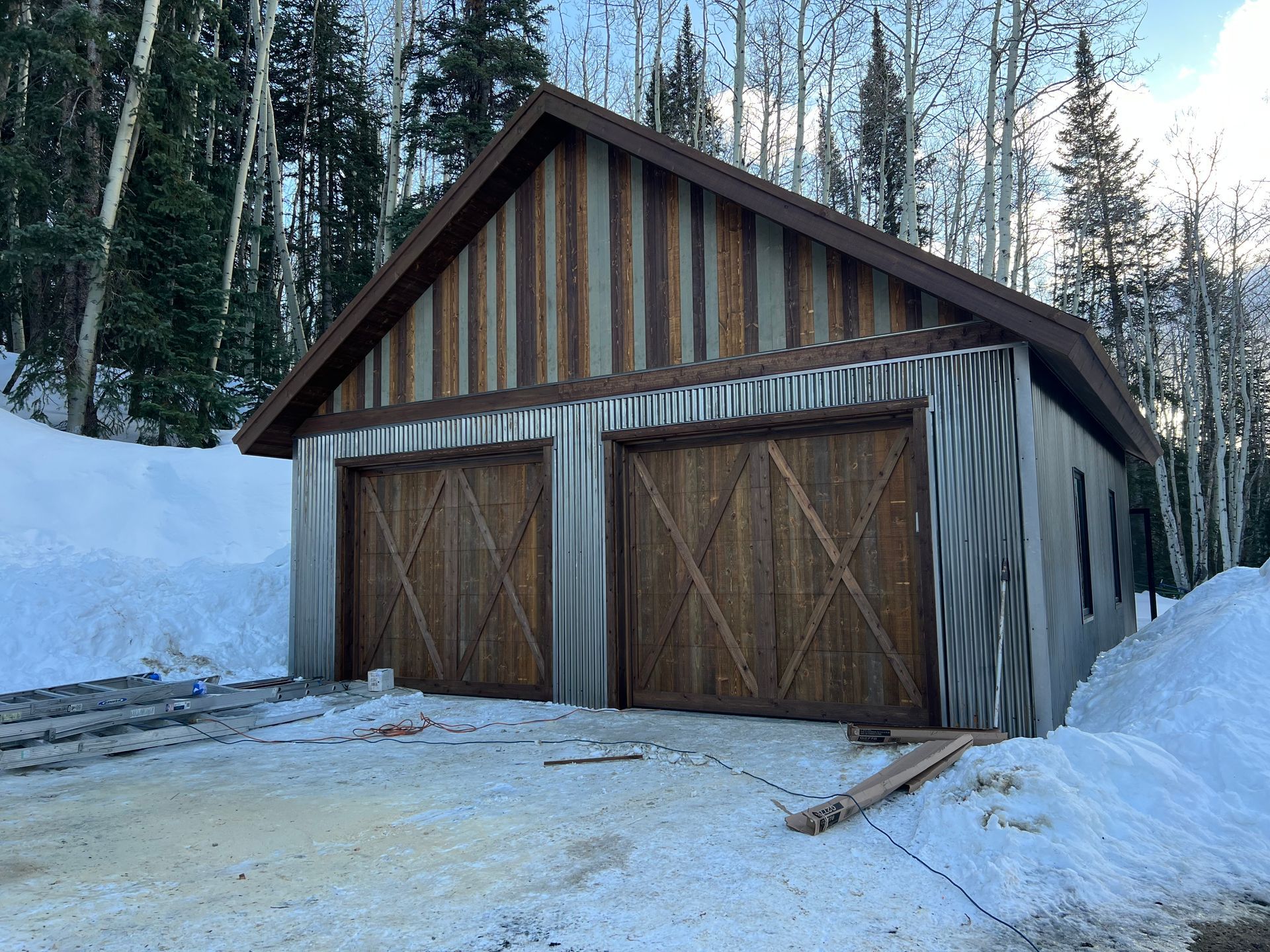 A garage with wooden doors is surrounded by snow and trees.