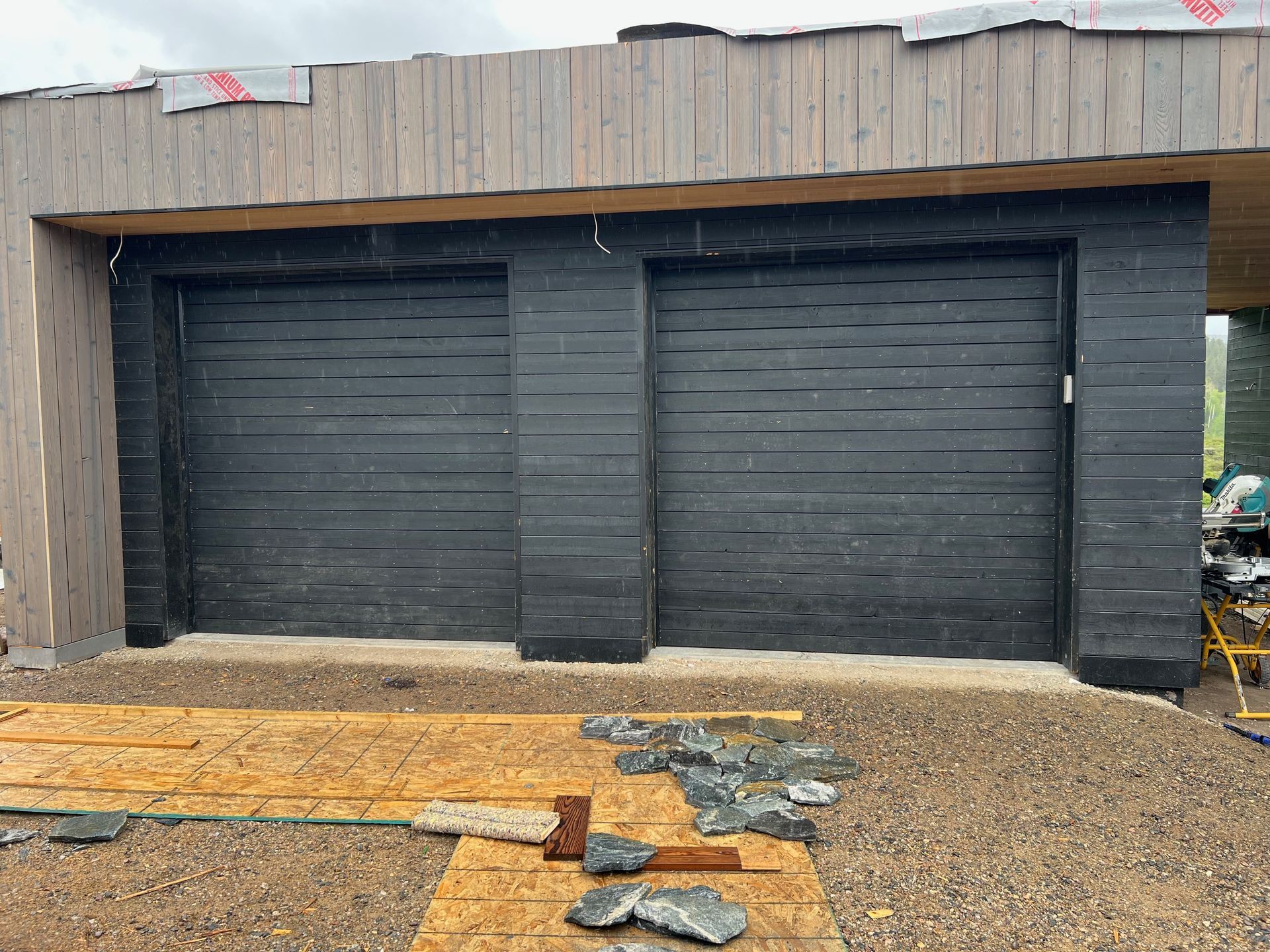 Two black garage doors in a building under construction with wood siding. Construction materials are scattered in front.